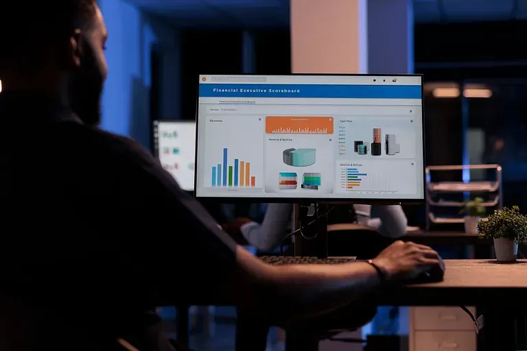 Un hombre con barba usa una computadora en una oficina oscura, viendo gráficos de datos en la pantalla.