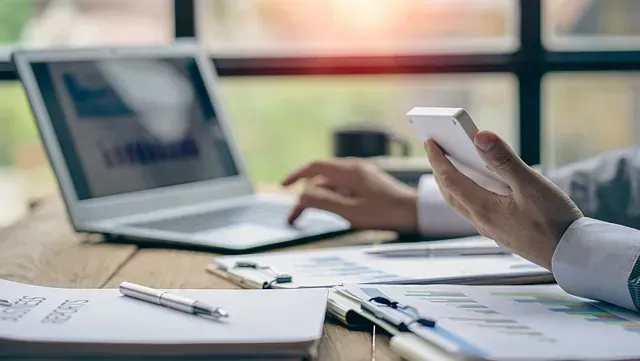 Una persona trabaja en un escritorio con una laptop y un teléfono inteligente, con documentos y gráficos frente a ella. La oficina cuenta con luz natural que entra por una ventana.