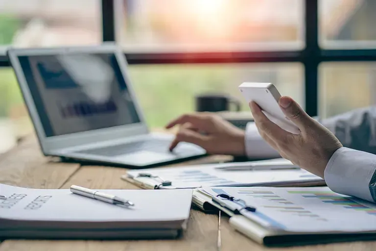 Persona trabajando en un escritorio con una computadora portátil, documentos y un teléfono móvil, bañada por la luz del sol.