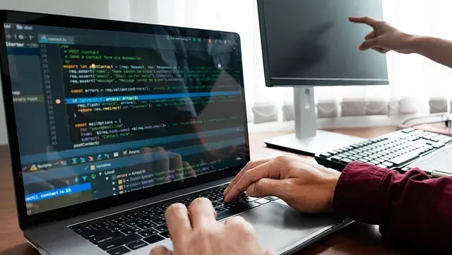 Persona programando en una laptop con un segundo monitor. Una mano escribe en el teclado y la otra apunta al monitor.