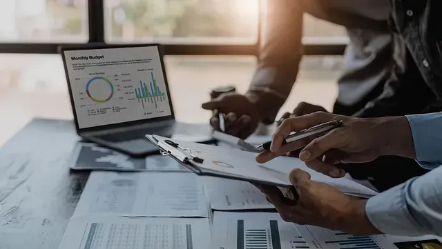 Dos personas revisando datos financieros, con una computadora portátil mostrando gráficos y documentos en un escritorio.