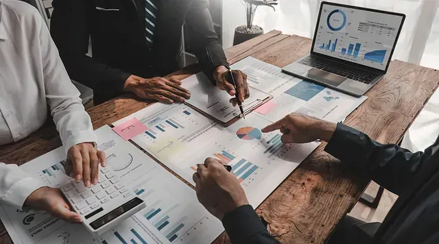 Tres personas analizan datos financieros alrededor de una mesa de madera con gráficos, una calculadora y una computadora portátil.