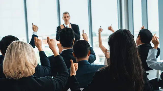Presentación empresarial. Los asistentes levantan la mano, en señal de aprobación, hacia un presentador en un moderno entorno de oficina con vistas panorámicas.