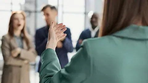 Una persona con una chaqueta verde hace un gesto hacia un grupo de colegas en una oficina.