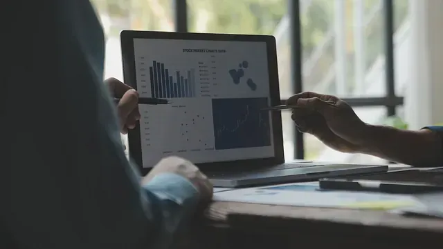 Dos personas revisan la pantalla de una computadora portátil con gráficos financieros, posiblemente en una oficina. Una de ellas señala la pantalla con un bolígrafo.