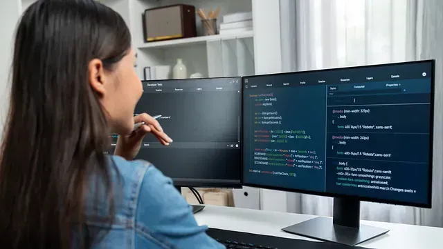 Mujer codificando en dos monitores en una oficina iluminada; el código se muestra en la pantalla derecha, sonriendo y señalando la pantalla.
