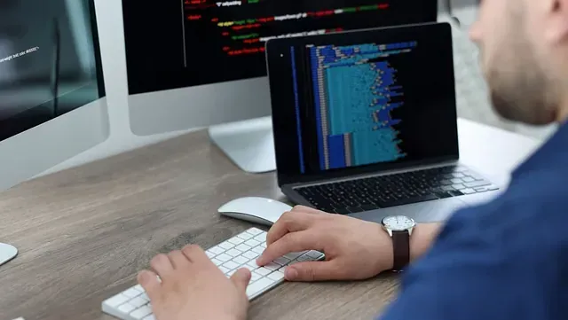 Persona escribiendo en un teclado frente a una laptop y dos monitores, probablemente codificando. Se muestra un código azul y blanco en las pantallas.