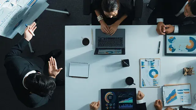 Vista aérea de una reunión de negocios: personas en una mesa blanca, examinando datos en gráficos y pantallas de computadoras portátiles, con un hombre gesticulando enfáticamente.