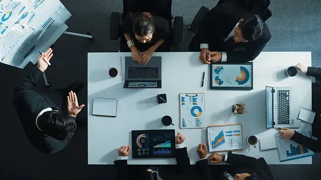 Equipo de negocios con traje en una mesa de conferencias blanca, revisando gráficos y discutiendo estrategias; toma cenital.