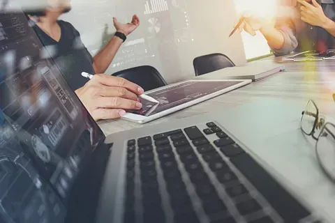 Personas en una reunión en una mesa con computadoras portátiles y tabletas, discutiendo visualizaciones de datos proyectadas en el fondo.