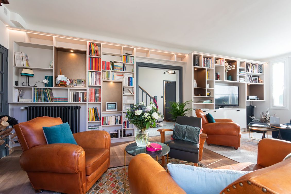 Salon avec bibliothèque intégrée, fauteuils en cuir et vue sur un escalier.