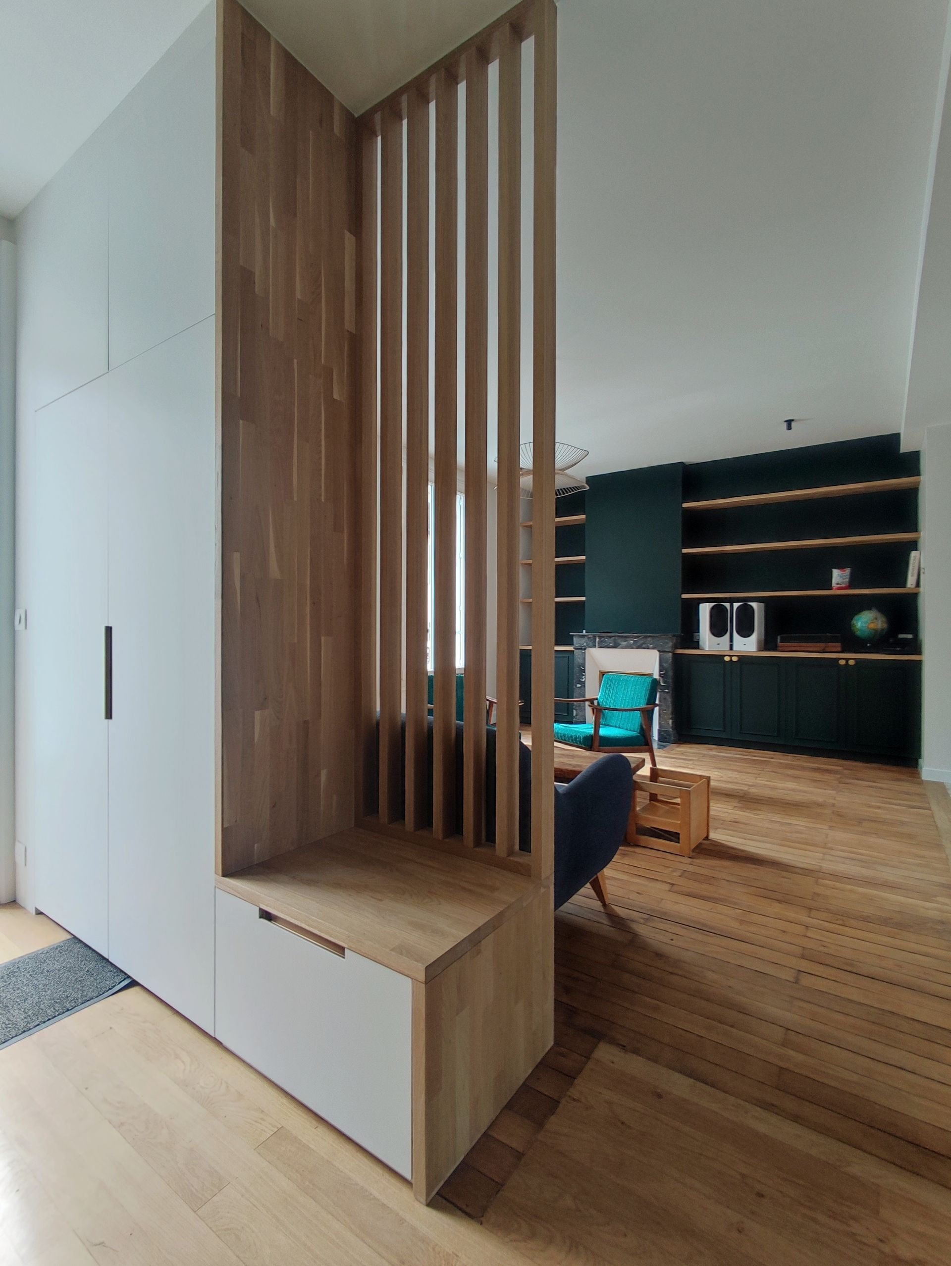 Paravent en bois à lattes verticales avec banquette intégrée, séparant l'entrée du salon avec parquet et mur vert foncé.