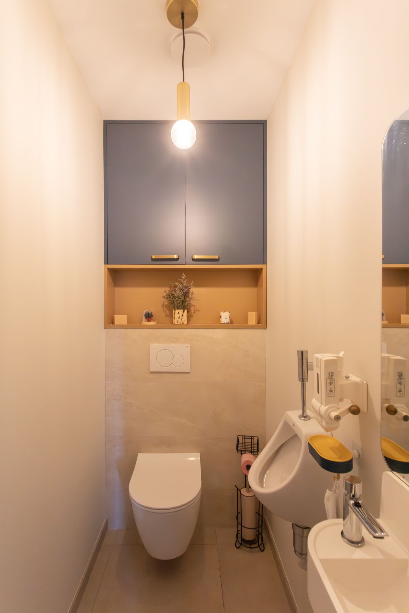 Des toilettes modernes et étroites avec WC, urinoir et petit lavabo. Meuble bleu au-dessus des toilettes.