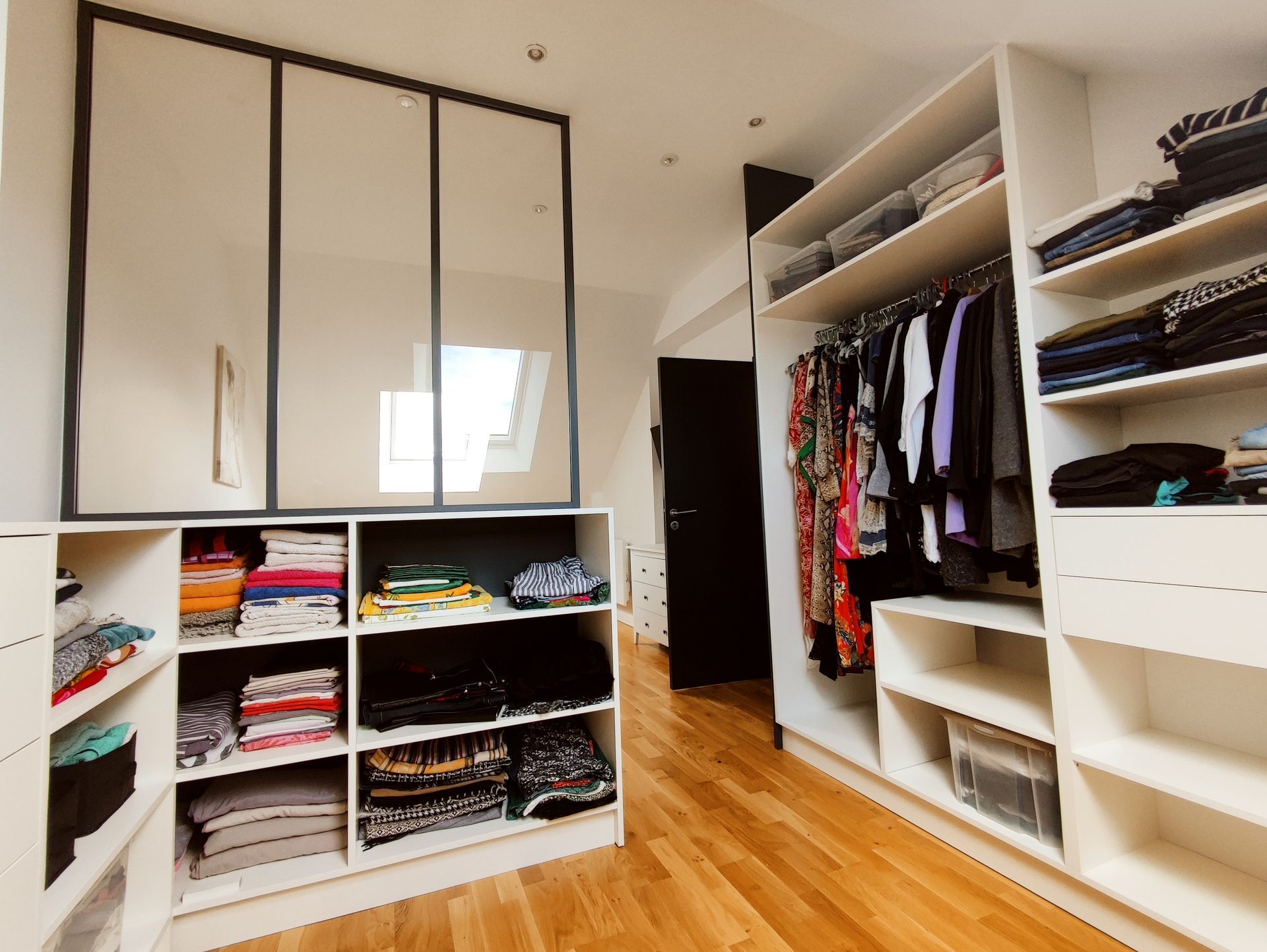 Dressing avec étagères et rangements blancs, vêtements et portes vitrées. Sol en bois et porte noire.
