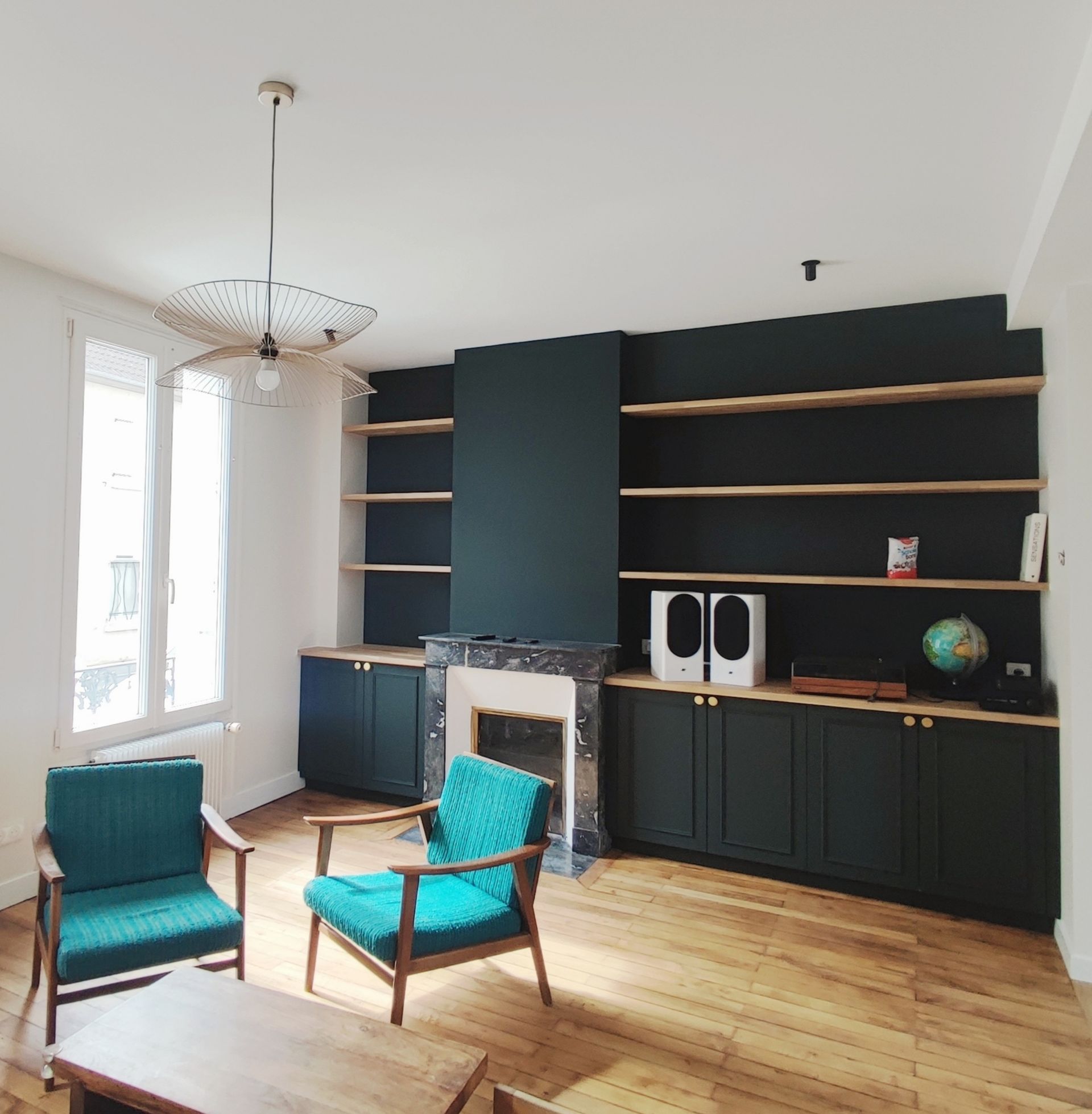 Salon avec fauteuils turquoise, meubles intégrés vert foncé, cheminée, parquet et suspension lumineuse.
