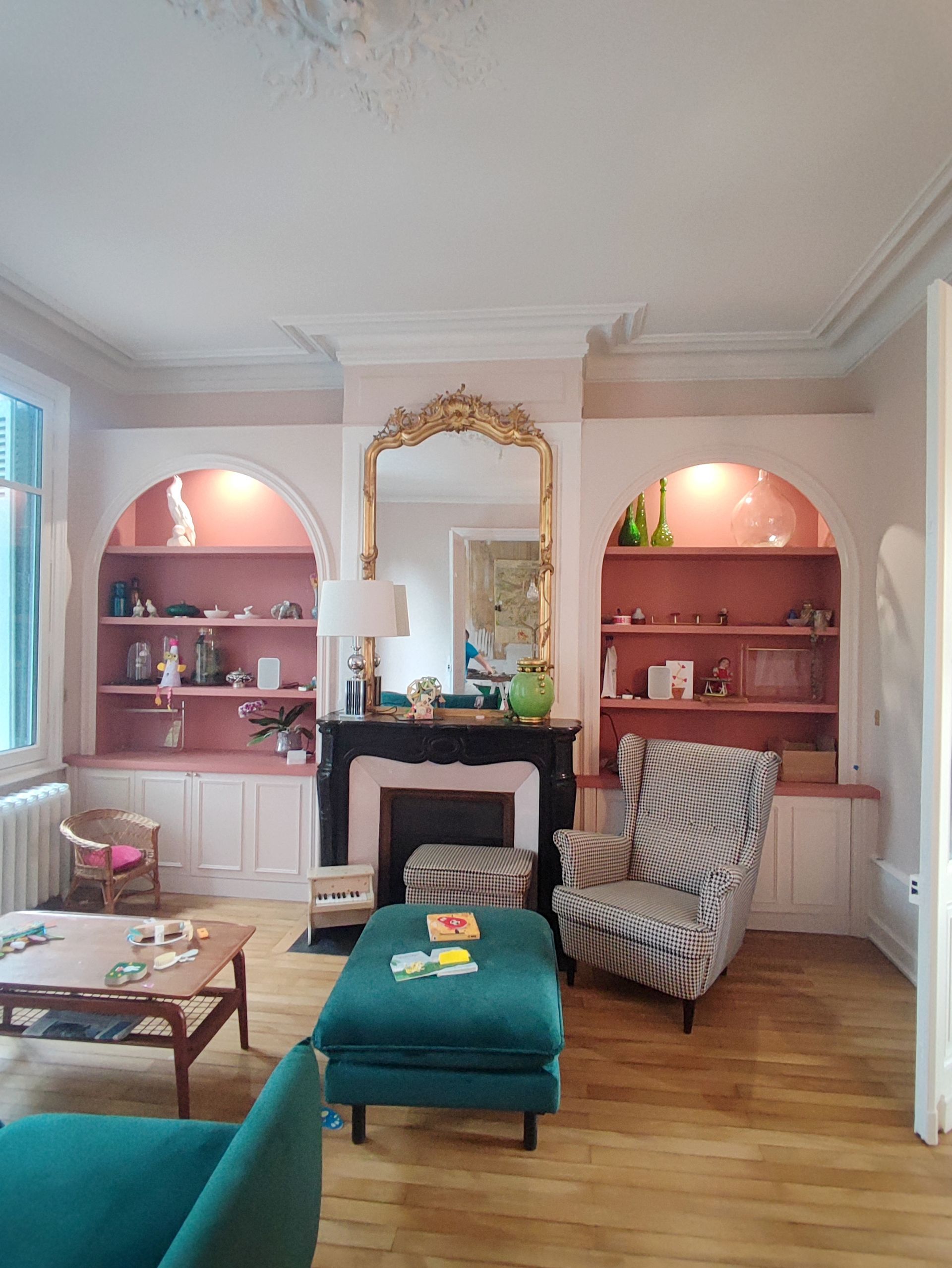 Salon avec touches de rose, cheminée, étagères intégrées, fauteuil et pouf turquoise.