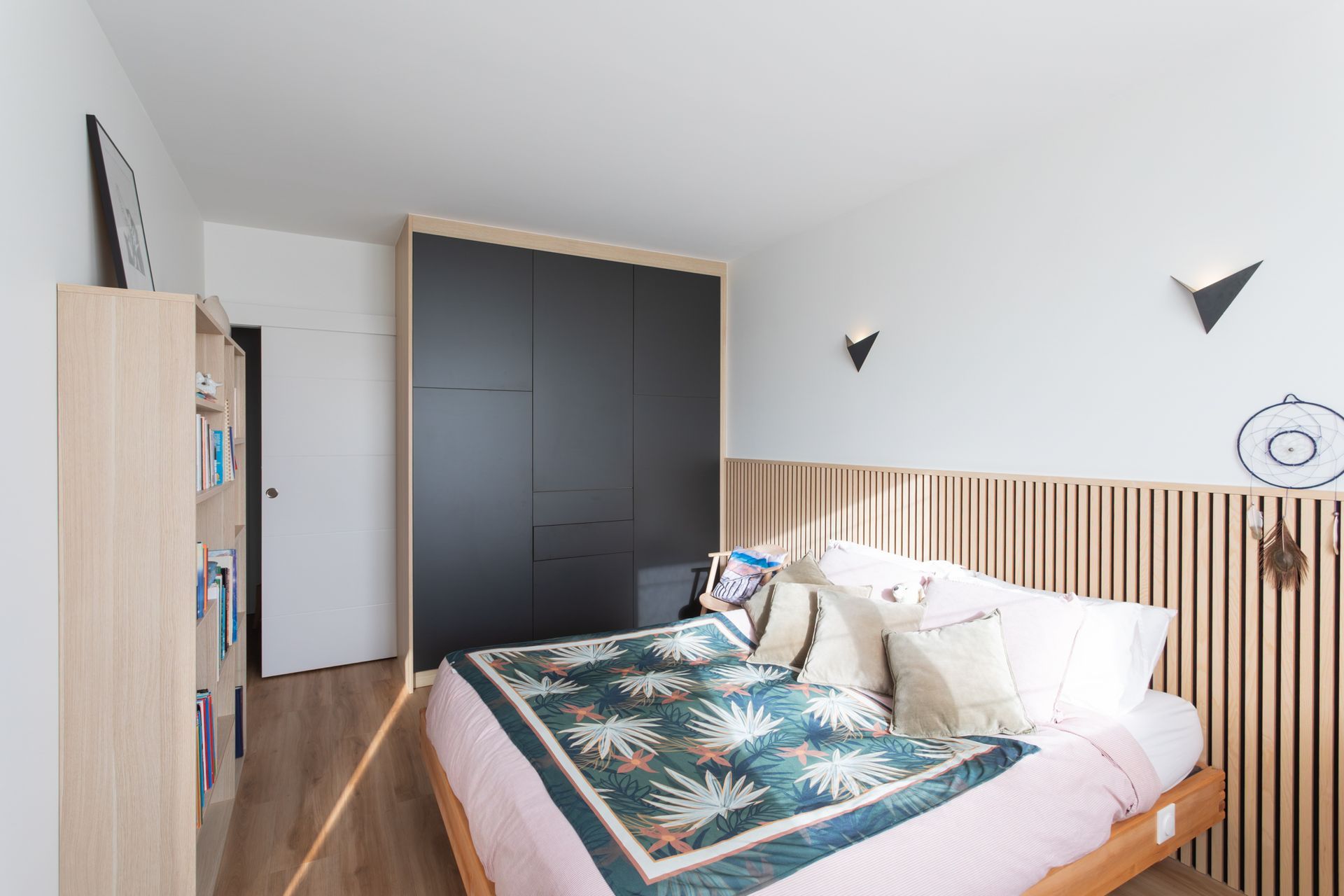 Chambre avec un lit en bois, une couverture à motifs, une armoire noire et un mur en bois clair.