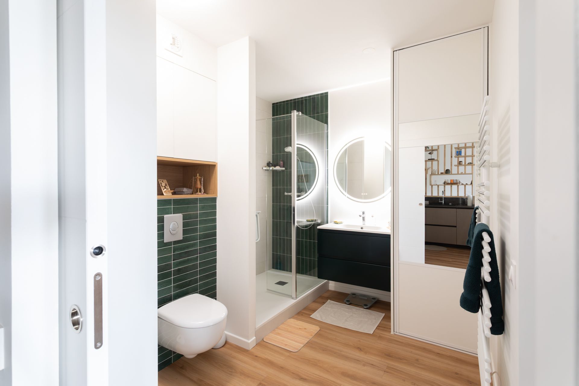 Salle de bains moderne avec douche carrelée verte, toilettes blanches, meuble-lavabo noir et deux miroirs ronds.
