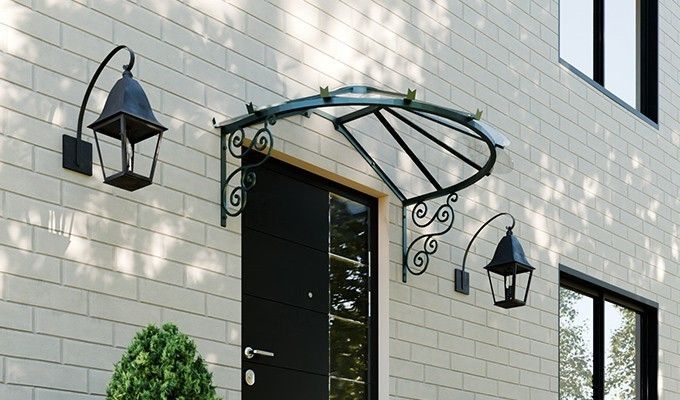 Extérieur d'une maison avec une porte noire, un auvent vert, deux lanternes murales et un mur en briques claires.
