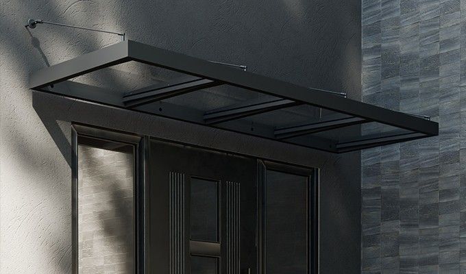 Un auvent noir recouvre une porte, avec des panneaux de verre et des bras de soutien, sur un bâtiment sombre.