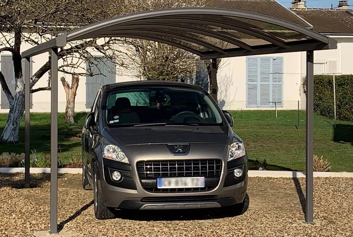 Un SUV gris est garé sous un abri métallique devant une maison.