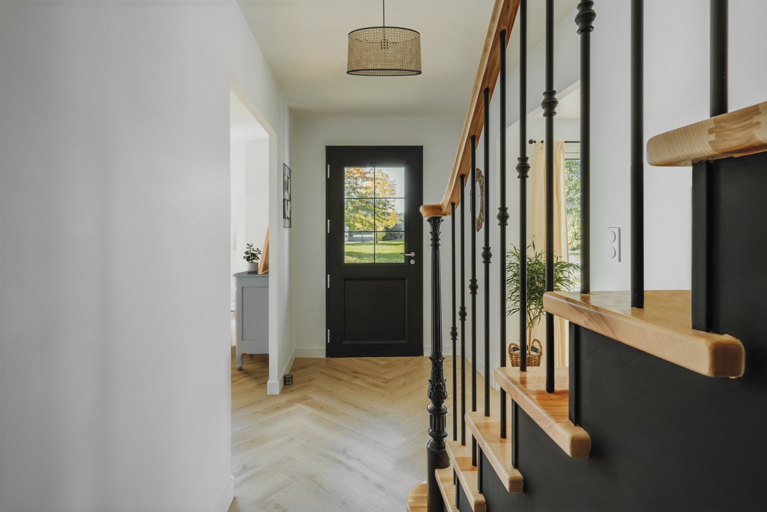 Couloir avec porte d'entrée noire, escalier en bois, rampe noire et parquet à chevrons.