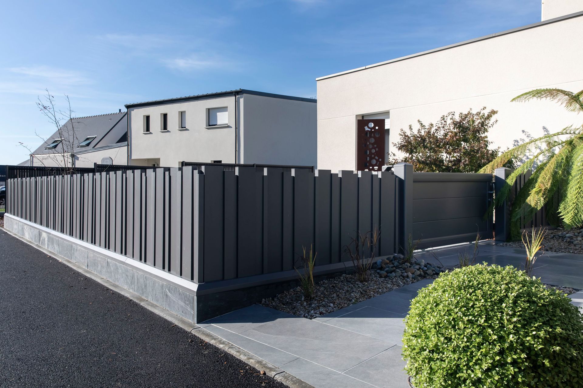 Clôture gris foncé bordant une maison et une rue sous un ciel bleu.