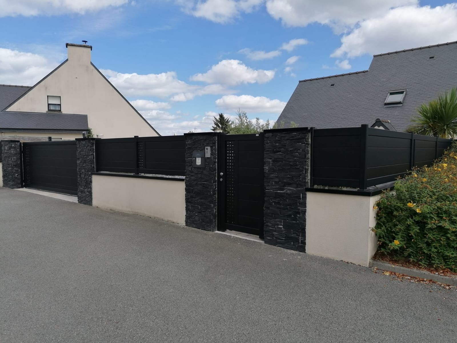 Clôture et portail noirs avec piliers en pierre devant une maison, dans une rue grise.