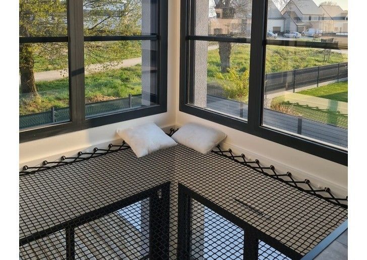 Coin intérieur avec fenêtres à cadre noir, filet de sécurité et coussins blancs. Vue sur un espace extérieur.