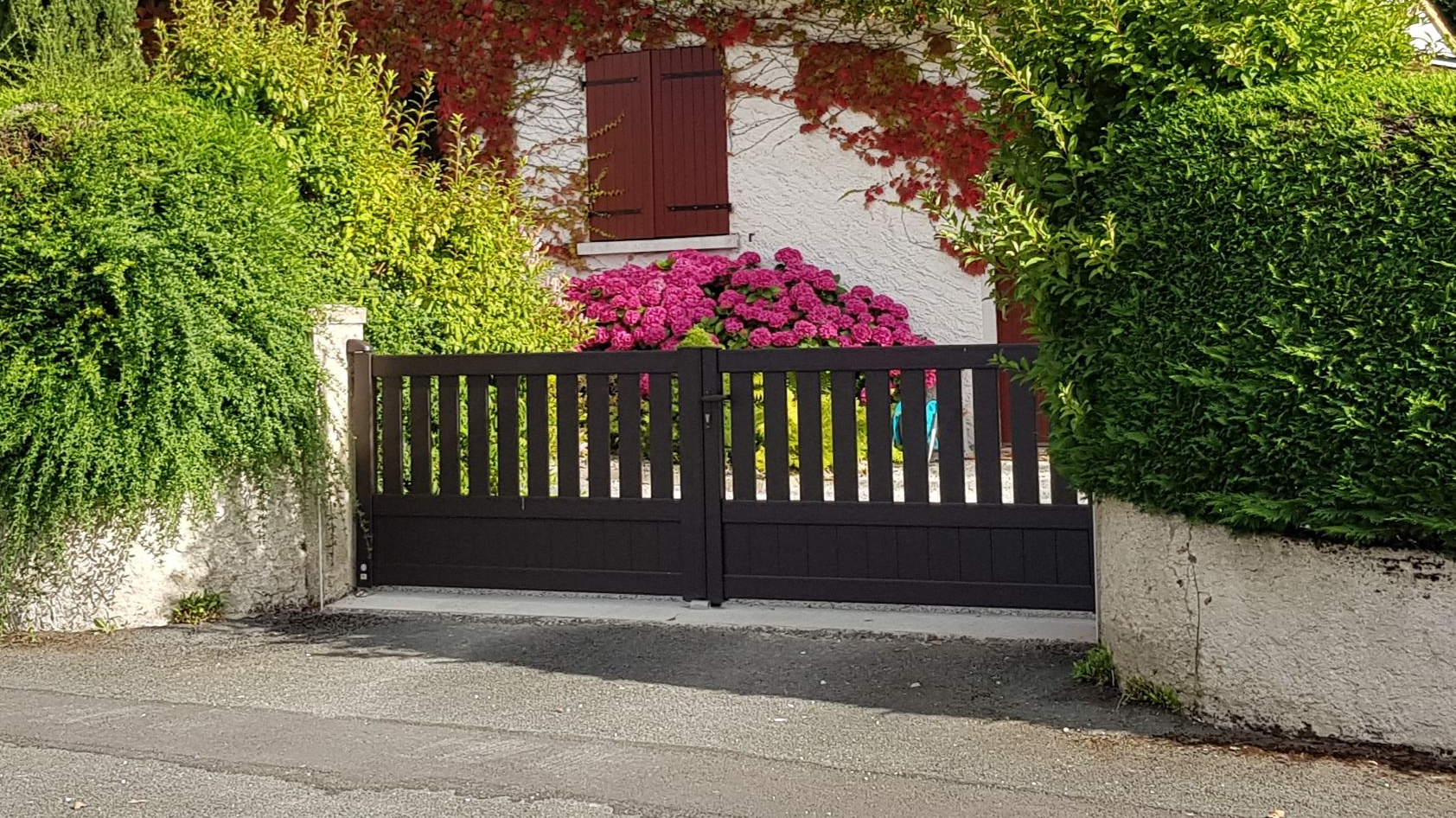 Un portail marron se dresse devant une maison blanche aux volets rouges.