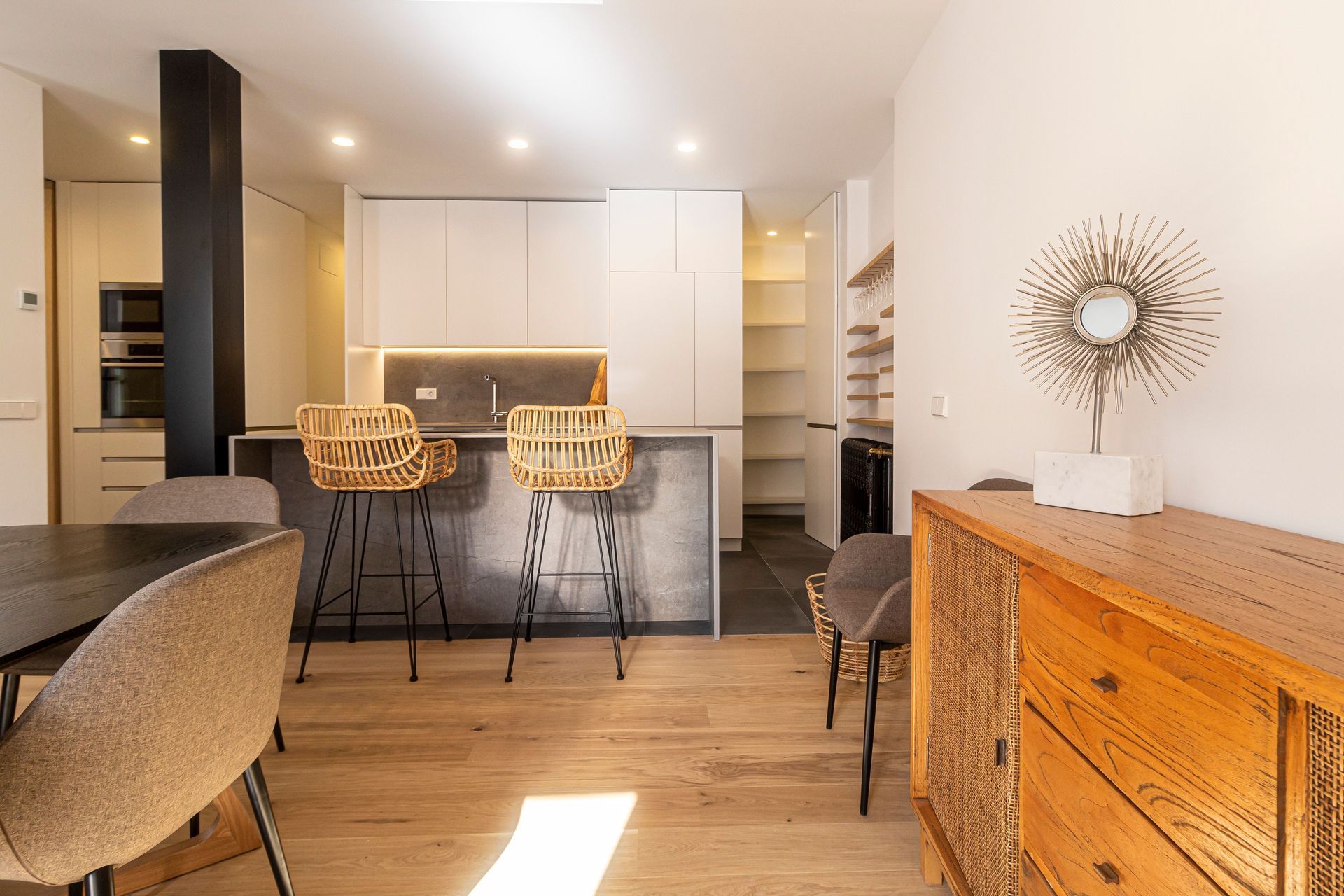 Cocina luminosa y moderna con isla, taburetes de bar de mimbre, armarios blancos, suelos de madera y un espejo en forma de sol radiante