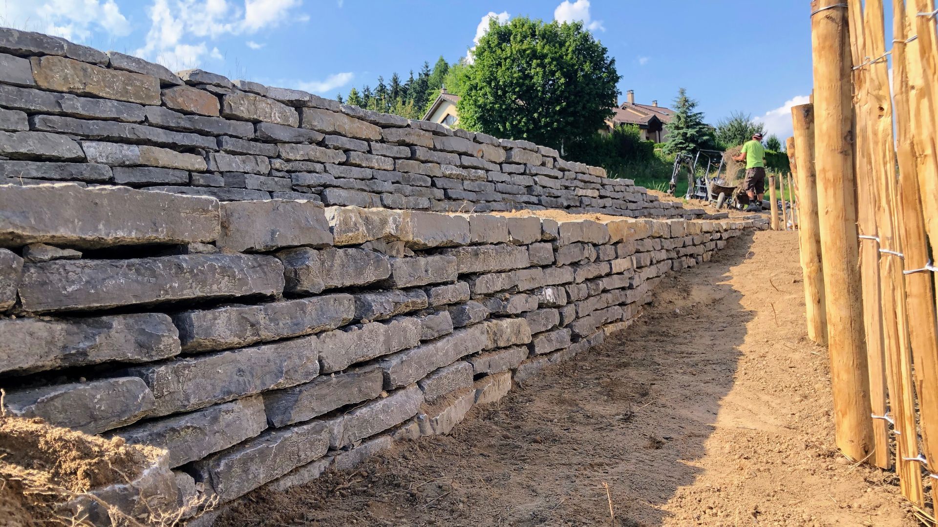 Mur de soutènement en pierre en construction; un chemin et des poteaux en bois sont visibles.
