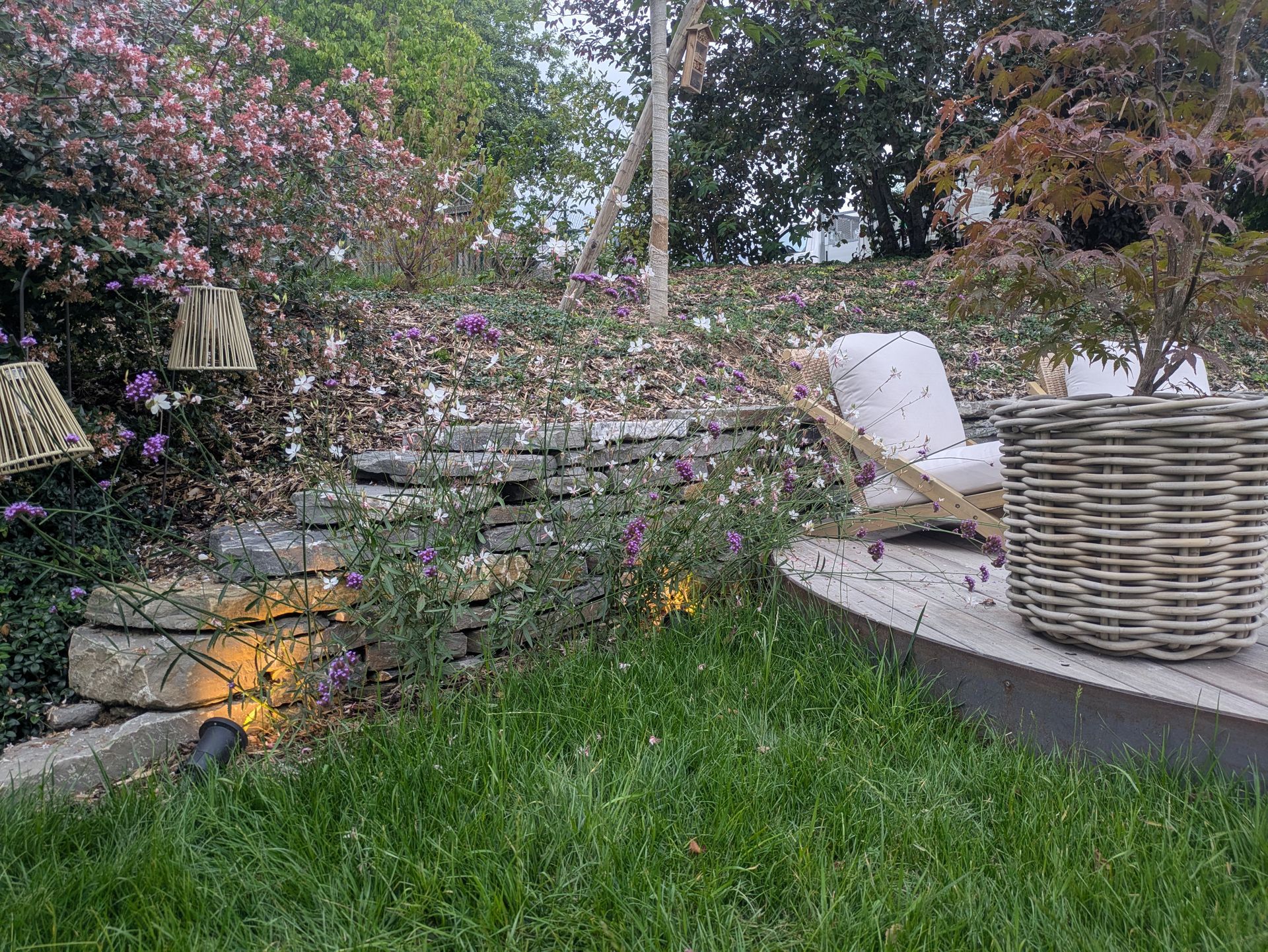 Mur en pierre et terrasse en bois dans un jardin avec sièges, verdure et lumières suspendues.