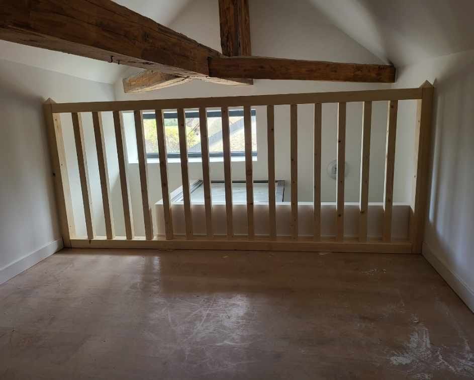 Une pièce avec une mezzanine à balustrade en bois donnant sur un espace inférieur, et présentant une grande poutre apparente en bois au plafond voûté.