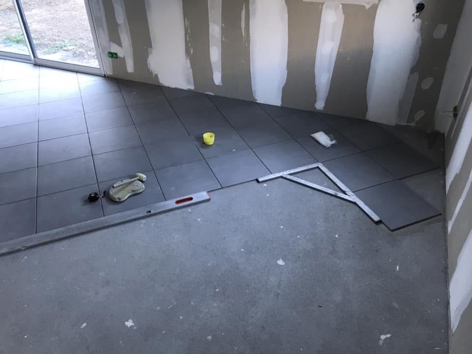 Carrelage gris partiellement posé dans une pièce aux murs en plaques de plâtre non finies, avec un outil de niveau et une équerre.