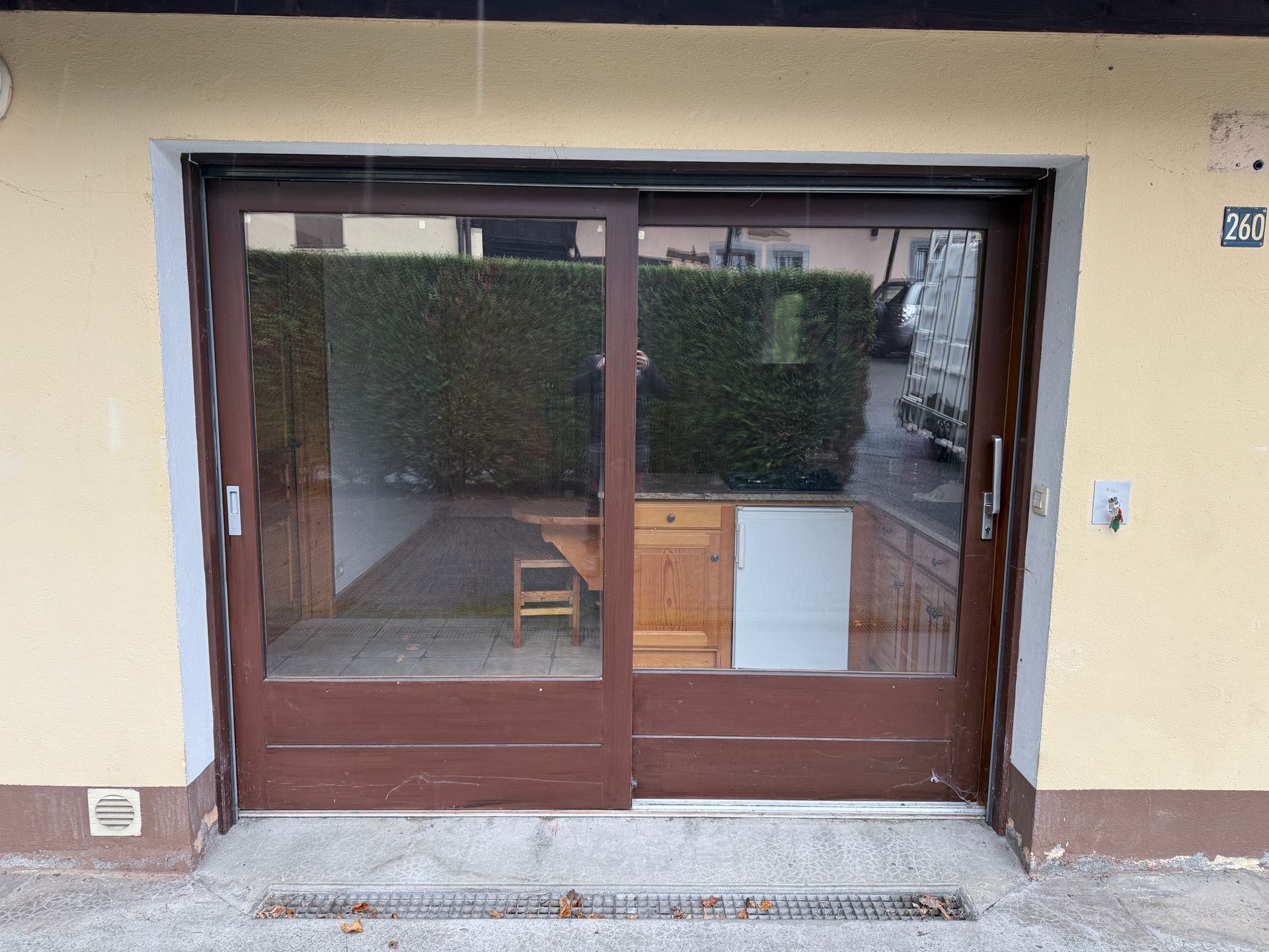 Porte coulissante en verre marron encastrée dans un mur extérieur beige, offrant une vue partielle sur une pièce et des haies à l'intérieur.