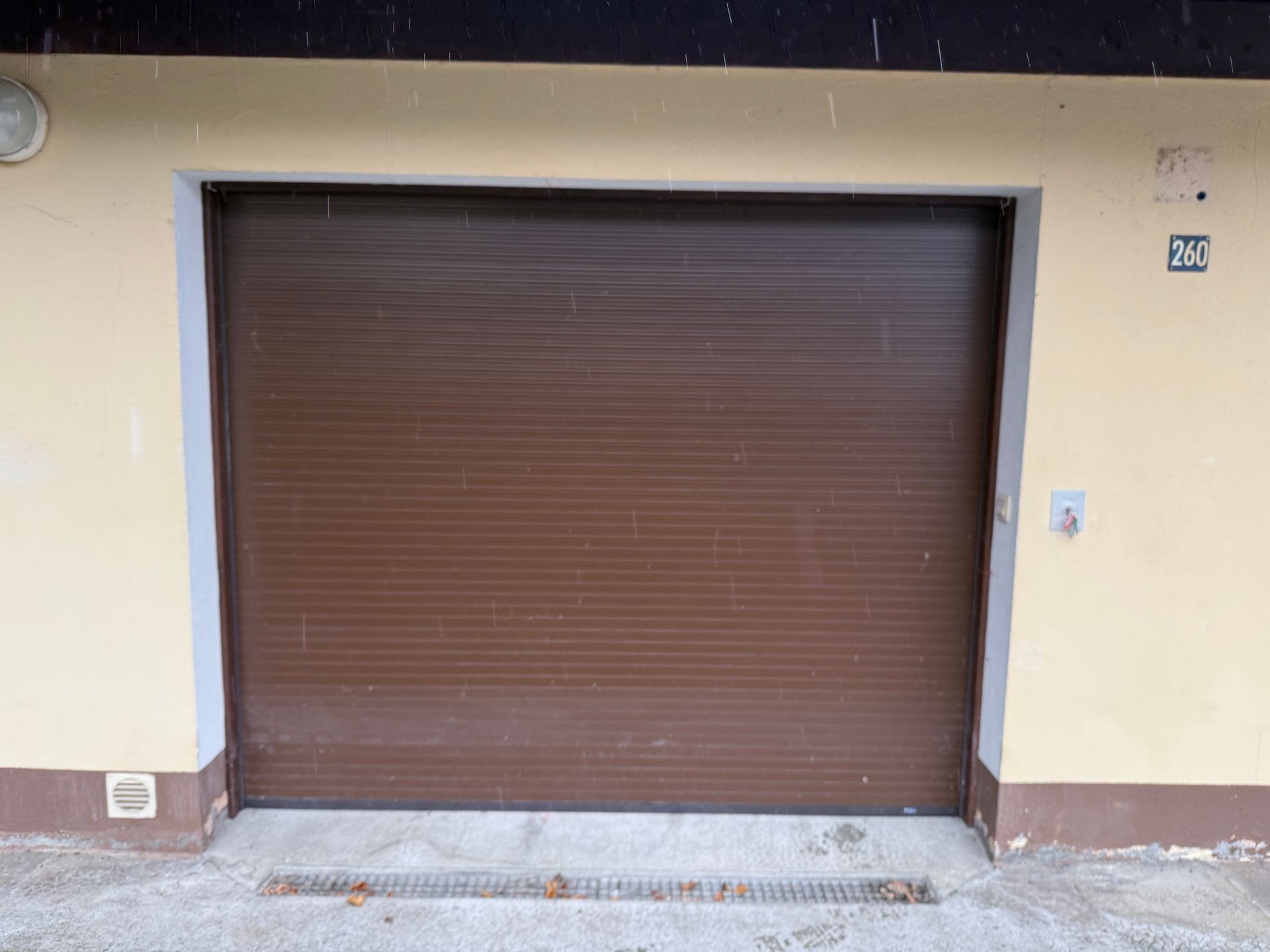 Une porte de garage enroulable brune à lames horizontales, centrée dans un mur beige, avec une allée en béton et un siphon de sol.