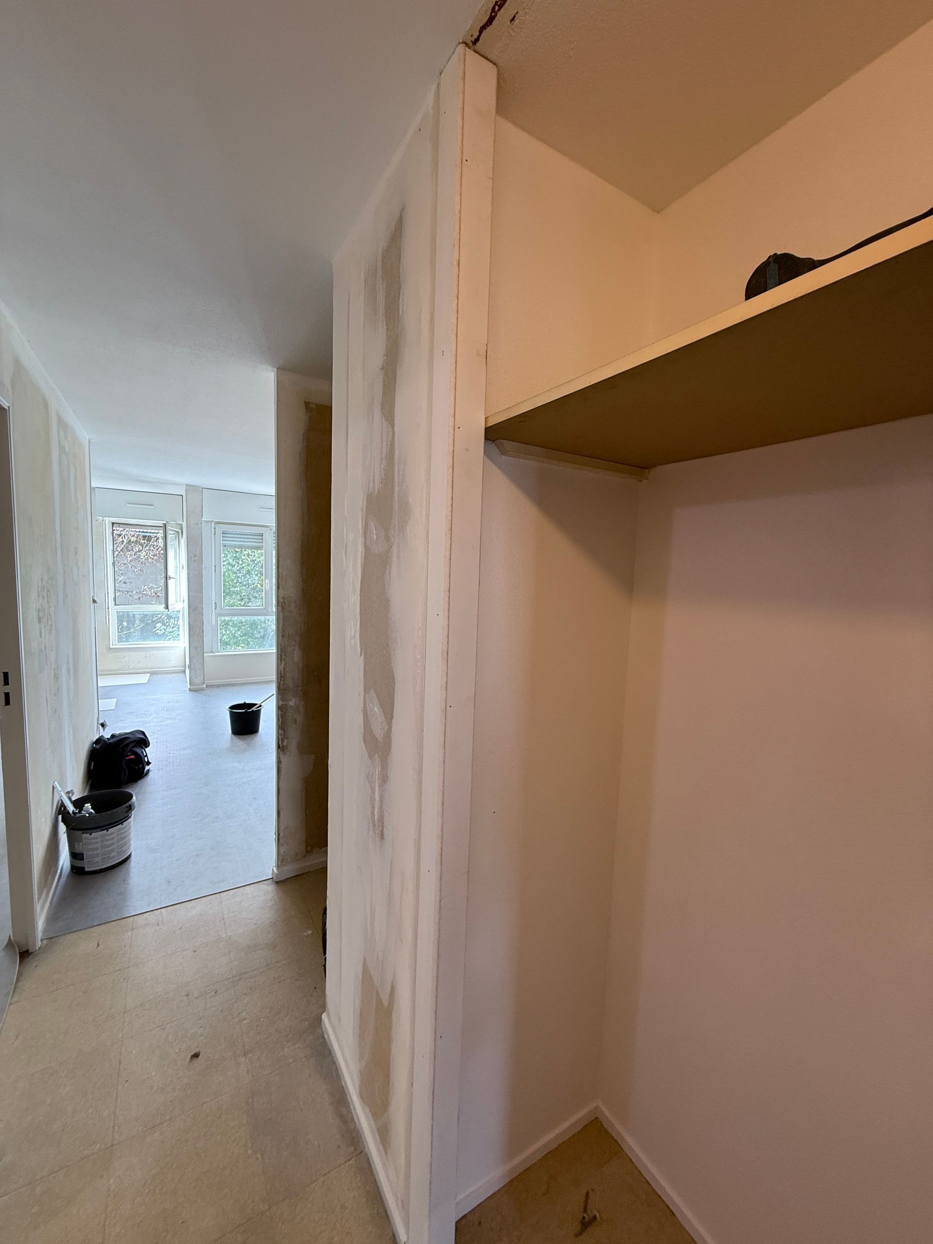 Une pièce en cours de rénovation avec un mur intérieur apparent, une étagère intégrée et un couloir menant à un espace vitré.