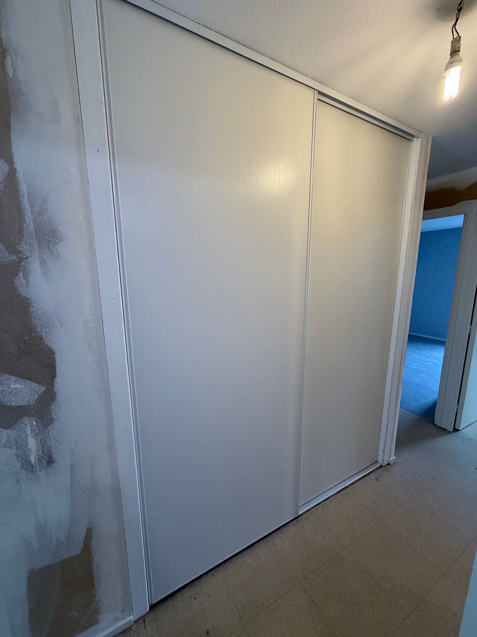 Deux portes de placard coulissantes blanches installées dans un couloir aux murs partiellement rebouchés et recouvert de moquette beige.