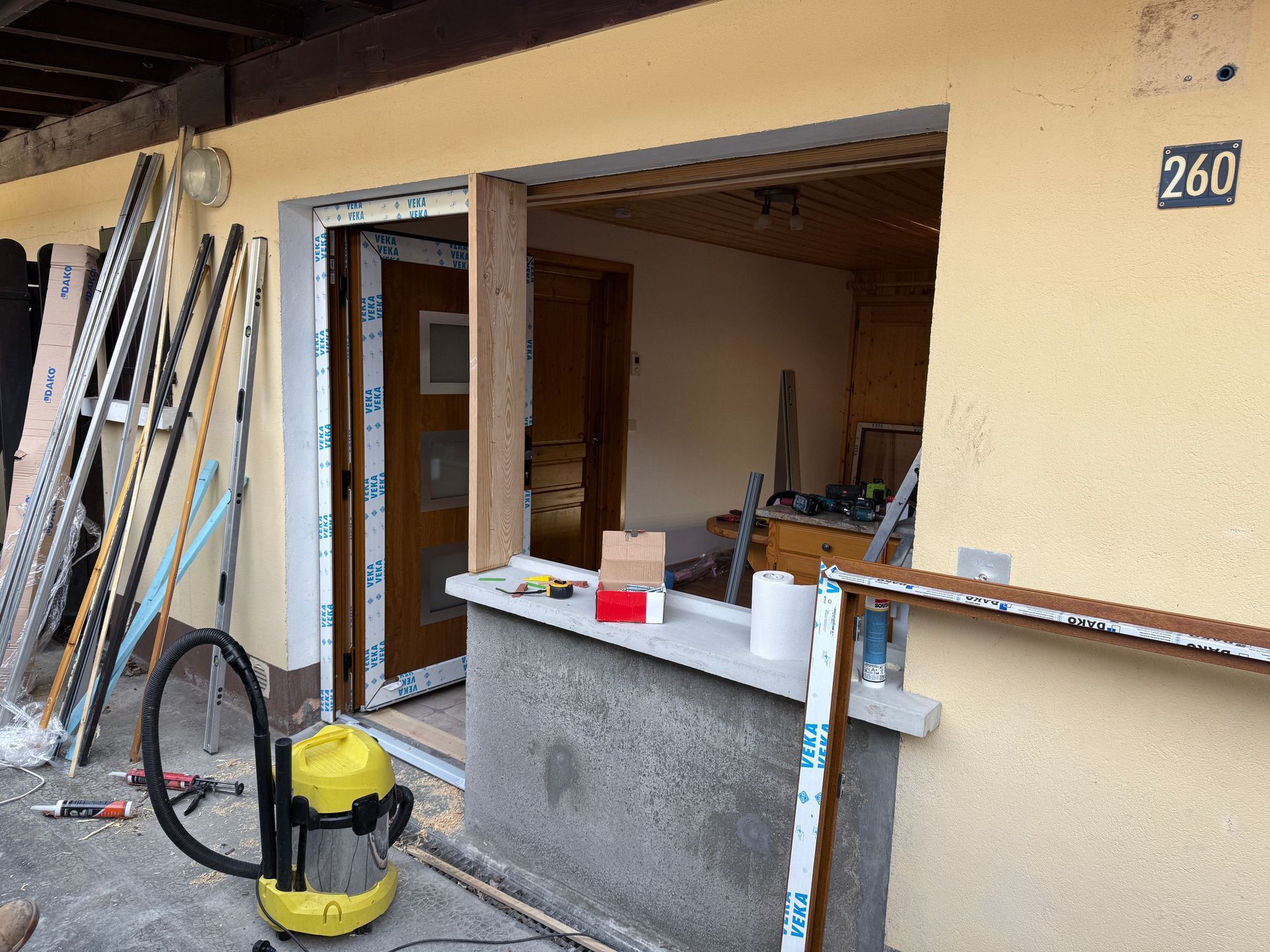 Un chantier inachevé, avec un nouveau cadre de fenêtre blanc en cours d'installation dans un mur en stuc jaune, à côté d'une porte.