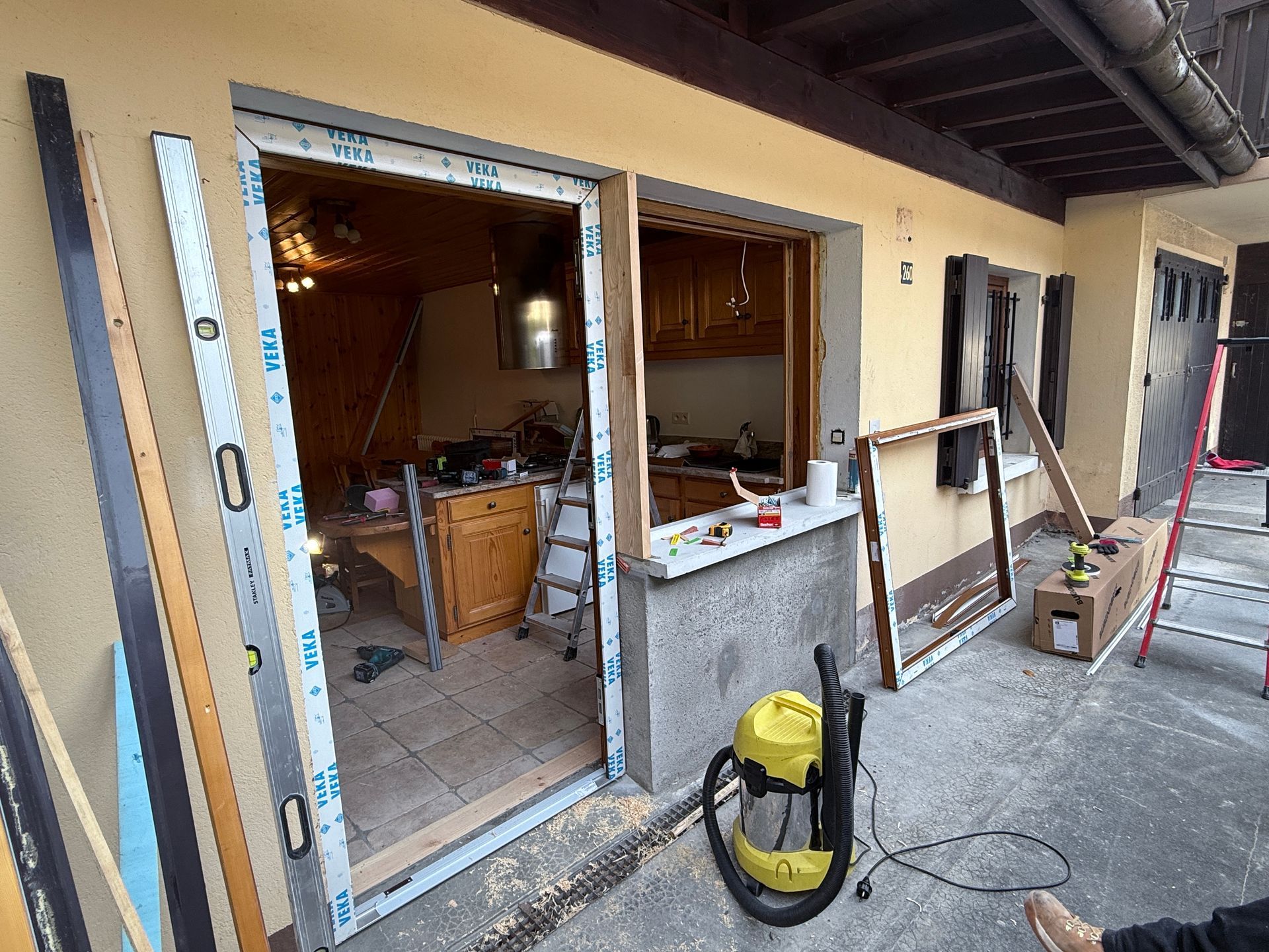 Un cadre de porte et une ouverture de fenêtre partiellement installés sur un mur extérieur beige, avec des outils et un aspirateur à proximité.