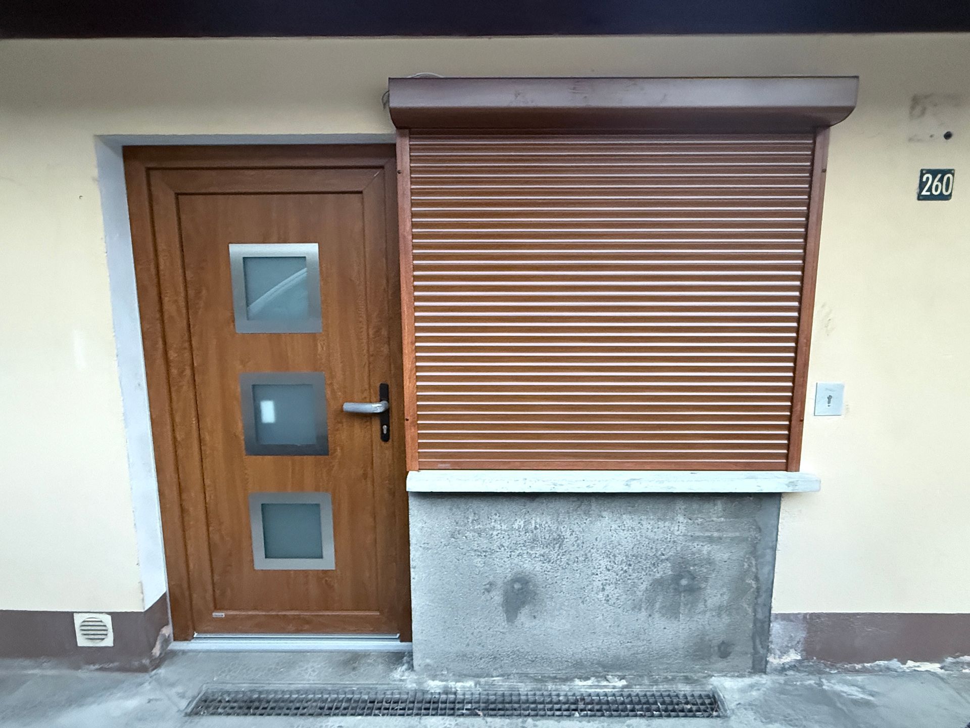 Une porte à motifs de bois avec trois fenêtres dépolies, à côté d'une fenêtre munie d'un volet roulant marron assorti, fermé.