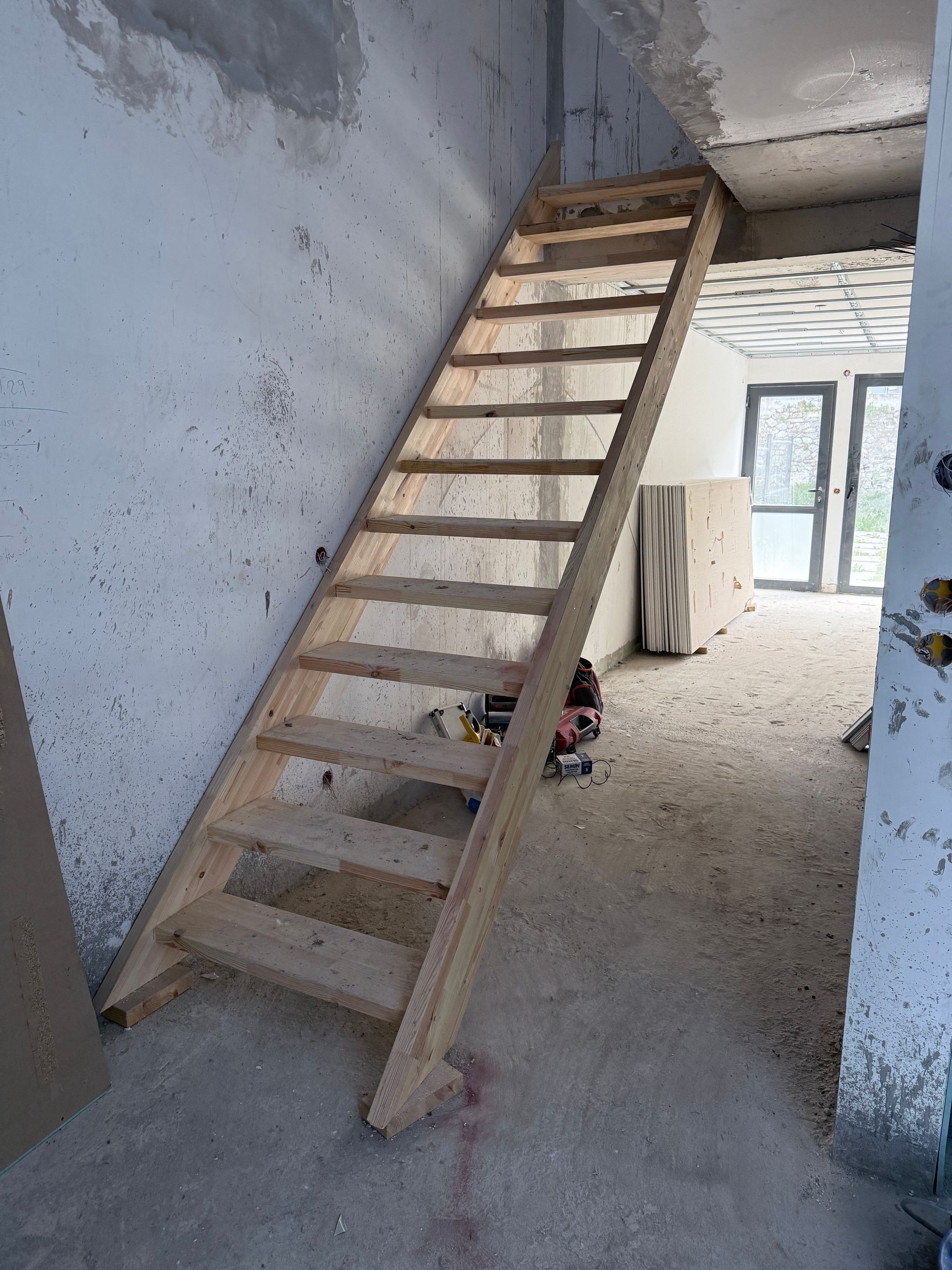Un escalier en bois raide et inachevé mène à un étage supérieur dans une pièce en construction aux murs et aux sols en béton gris.