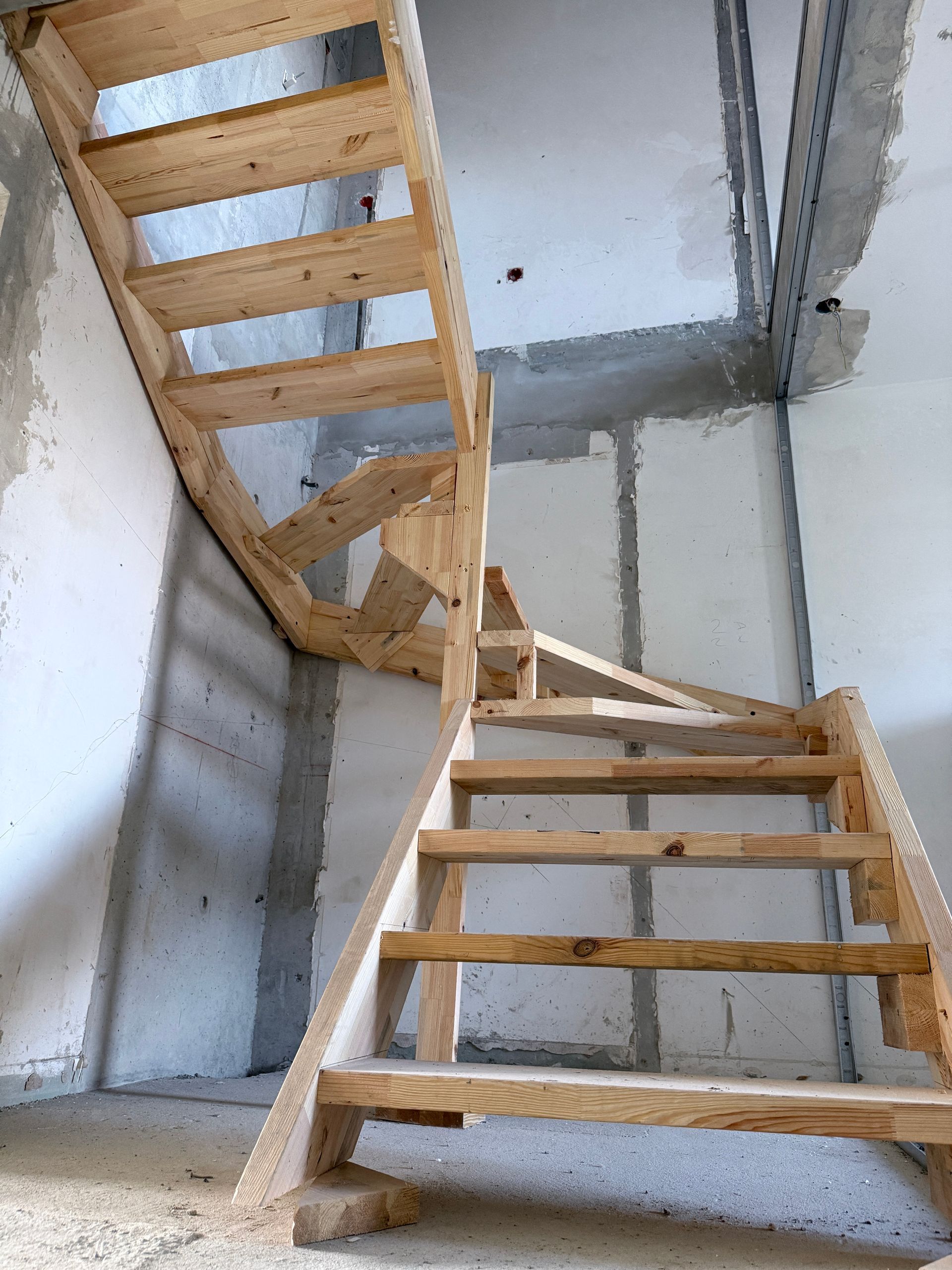 Un escalier en bois en construction dans une pièce aux murs blancs inachevés et à la charpente métallique apparente.