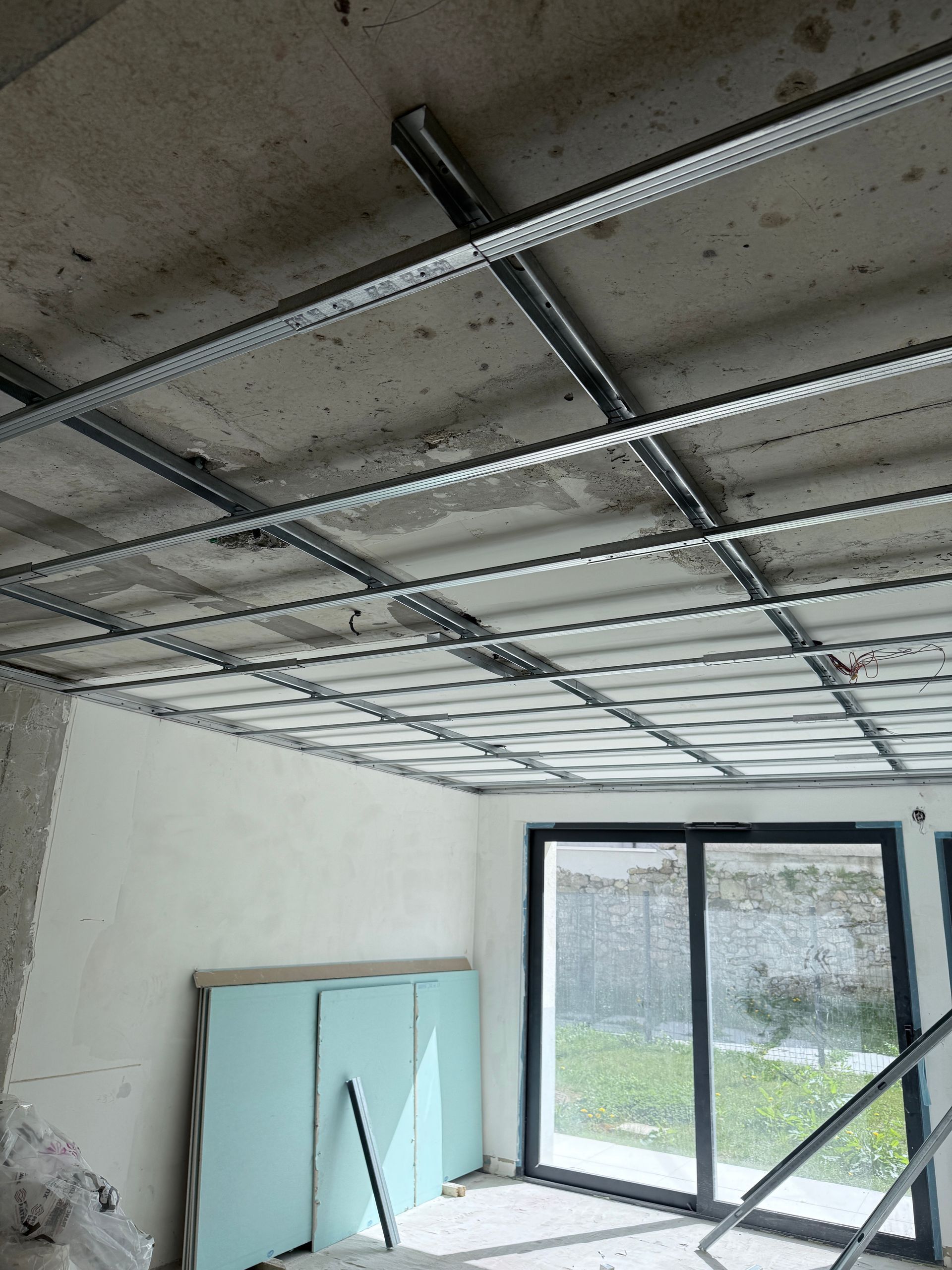 Une pièce en construction avec une grille de plafond métallique installée et des panneaux de plaques de plâtre appuyés contre le mur.