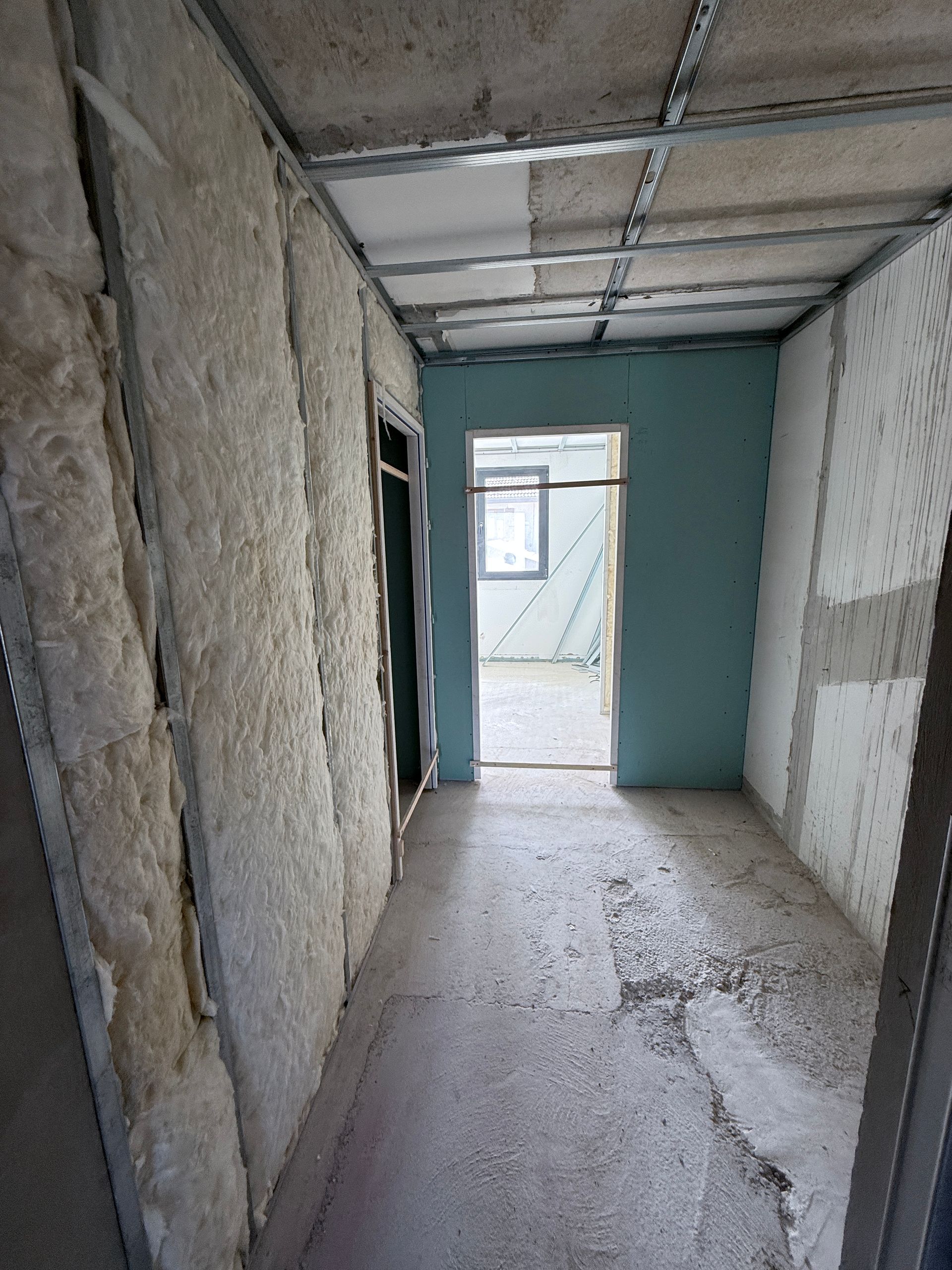 Un couloir de chantier avec une structure métallique, une isolation apparente et un mur peint en bleu menant à une porte ouverte.