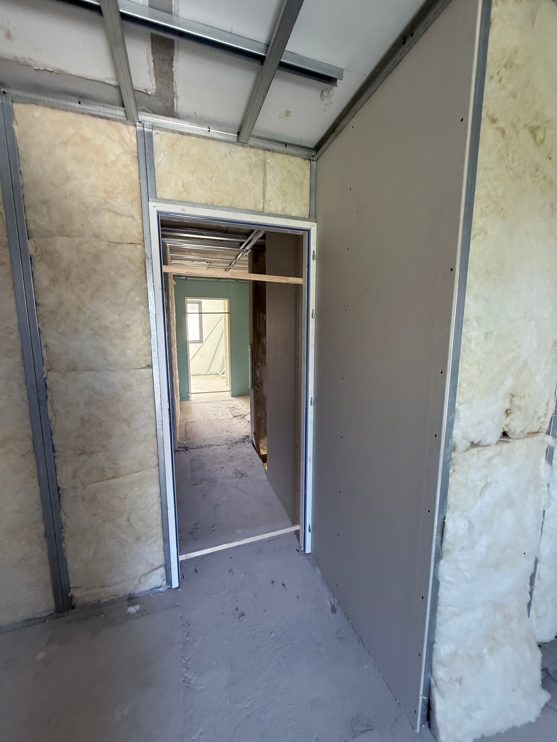 Une pièce inachevée avec des montants muraux métalliques, une isolation apparente et une porte donnant sur un autre espace cloisonné.