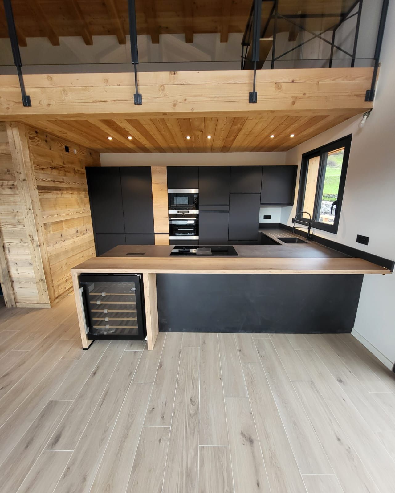 Cuisine moderne avec armoires noires mates, îlot central avec comptoir en bois, cave à vin intégrée et plafond en bois rustique.