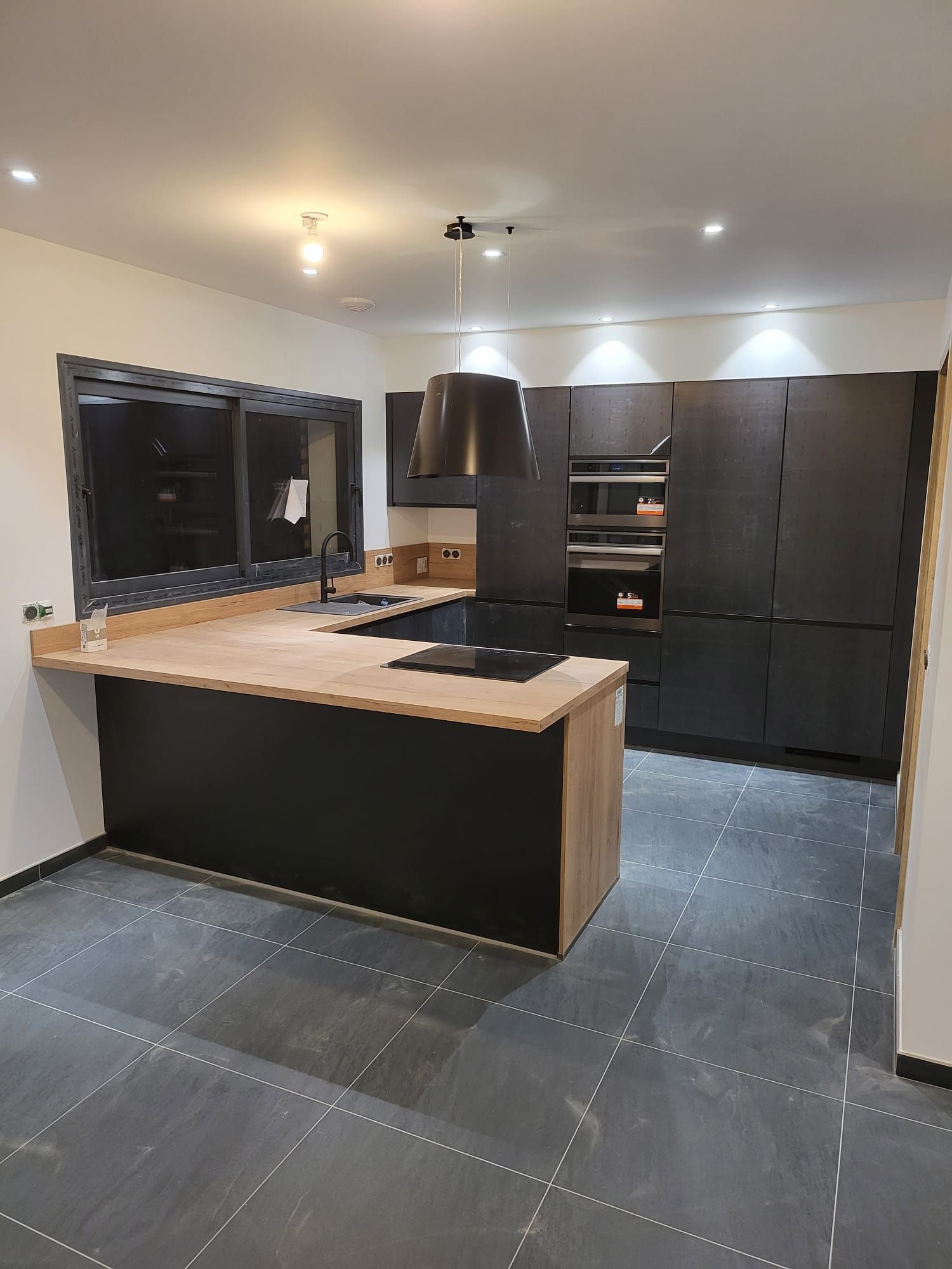 Cuisine moderne avec placards foncés, îlot de cuisine en bois clair, hotte suspendue et sol carrelé gris.