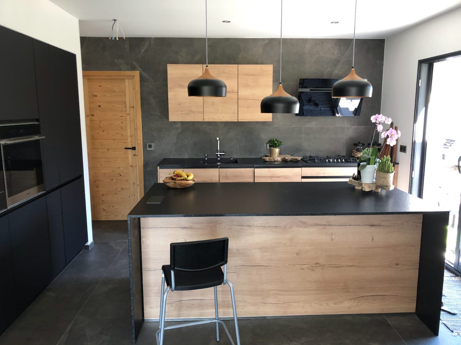 Une cuisine moderne comprend un îlot central noir avec un socle en bois, des meubles de cuisine noirs assortis et des placards muraux en bois clair.